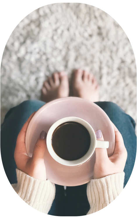 woman wearing cozy sweater rests barefoot on a lush rug holding a cup of coffee in a mug on her lap